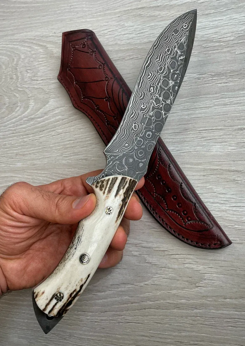 Un cuțit Bowie autentic de Damasc, cu m&acirc;ner din corn de cerb și arțar, forjat &icirc;n 150 de straturi, lucrat manual de un fierar.