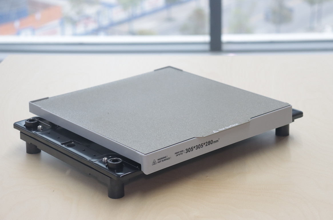3D 打印机热床特写，表面带纹理并标注尺寸(305&times;305&times;280mm&sup2;)，置于木桌上。作为 CoreXY 及其他 3D 打印机支撑打印物体的核心组件。