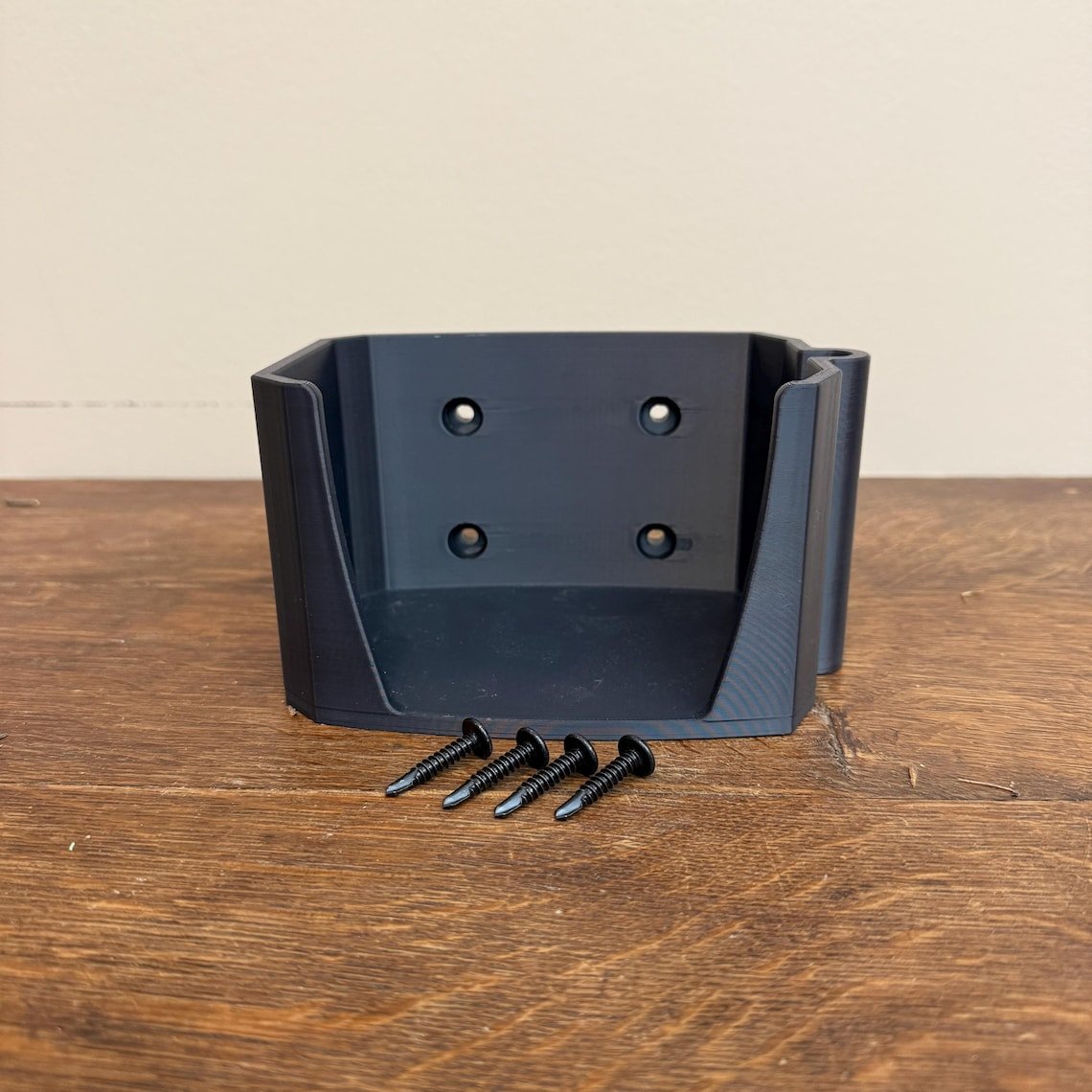 May include: A black plastic wall mount with four screw holes and a small lip on the side. Four black screws are in front of the mount. May include: A black plastic wall mount with four screw holes and a small lip on the side. Four black screws are in front of the mount.
