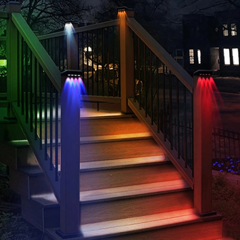 Four solar-powered post lights with color-changing LEDs illuminate a wooden staircase with railings at night. The lights emit white, blue, orange, and red beams downwards onto the steps and posts. A house with illuminated windows is visible in the background.