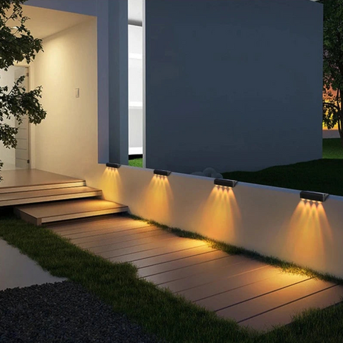 Four solar-powered fence lights are mounted along a low wall, illuminating a wooden walkway at dusk or night. The lights cast beams downwards onto the path. A modern house with warm interior lighting and a dark exterior wall is visible in the background, along with a patch of grass.