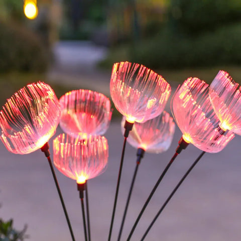 Several solar-powered garden lights with a translucent, jellyfish-like shape emit a soft pink glow at dusk or night. They are on thin dark stems and are seen outdoors with a blurred background.