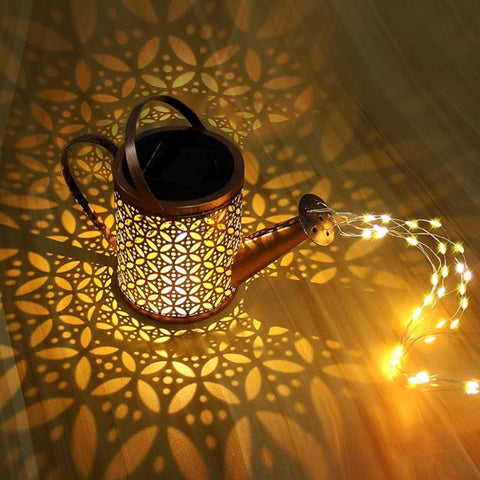 A decorative, bronze-toned watering can with intricate cutouts is illuminated from within, casting floral patterns of light onto a dark wooden surface. A string of small, warm-toned LED lights extends from the watering can's spout, creating the illusion of pouring light. The scene highlights a solar-powered garden decoration emitting a warm glow.