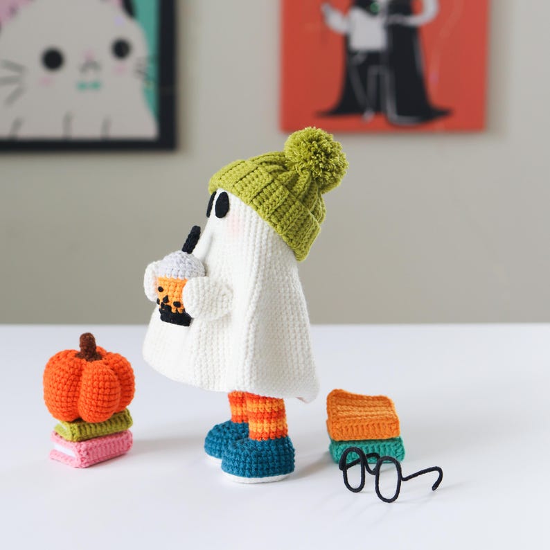 Cute crochet ghost doll with black glasses, green knit beanie, and blue-orange striped boots, sipping a boba tea. Displayed with a stack of colorful crochet books and an orange amigurumi pumpkin. Perfect handmade Halloween or fall decor. This is a PDF digital download crochet pattern to make your own cozy ghost plush and accessories
