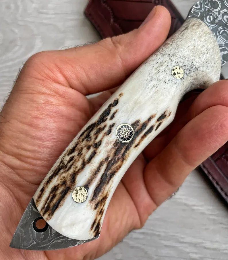 Un cuțit Bowie autentic de Damasc, cu m&acirc;ner din corn de cerb și arțar, forjat &icirc;n 150 de straturi, lucrat manual de un fierar.
