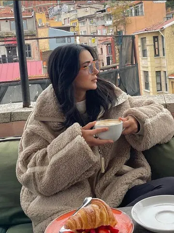 Person in einer Lammwolljacke hÃ¤lt eine Kaffeetasse, auf einem Teller liegt ein Croissant und Erdbeeren vor einer farbenfrohen Stadtkulisse.