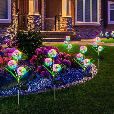 Several solar-powered garden lights that resemble stylized dandelions. They are placed among flowers and greenery in a front yard at dusk or dawn, casting a soft, multicolored glow. The house's facade is visible in the background.