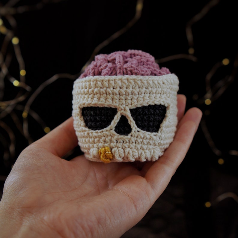 May include: A crocheted skull-shaped container with a pink crocheted brain inside. The skull is white with black eye sockets and a yellow button nose.