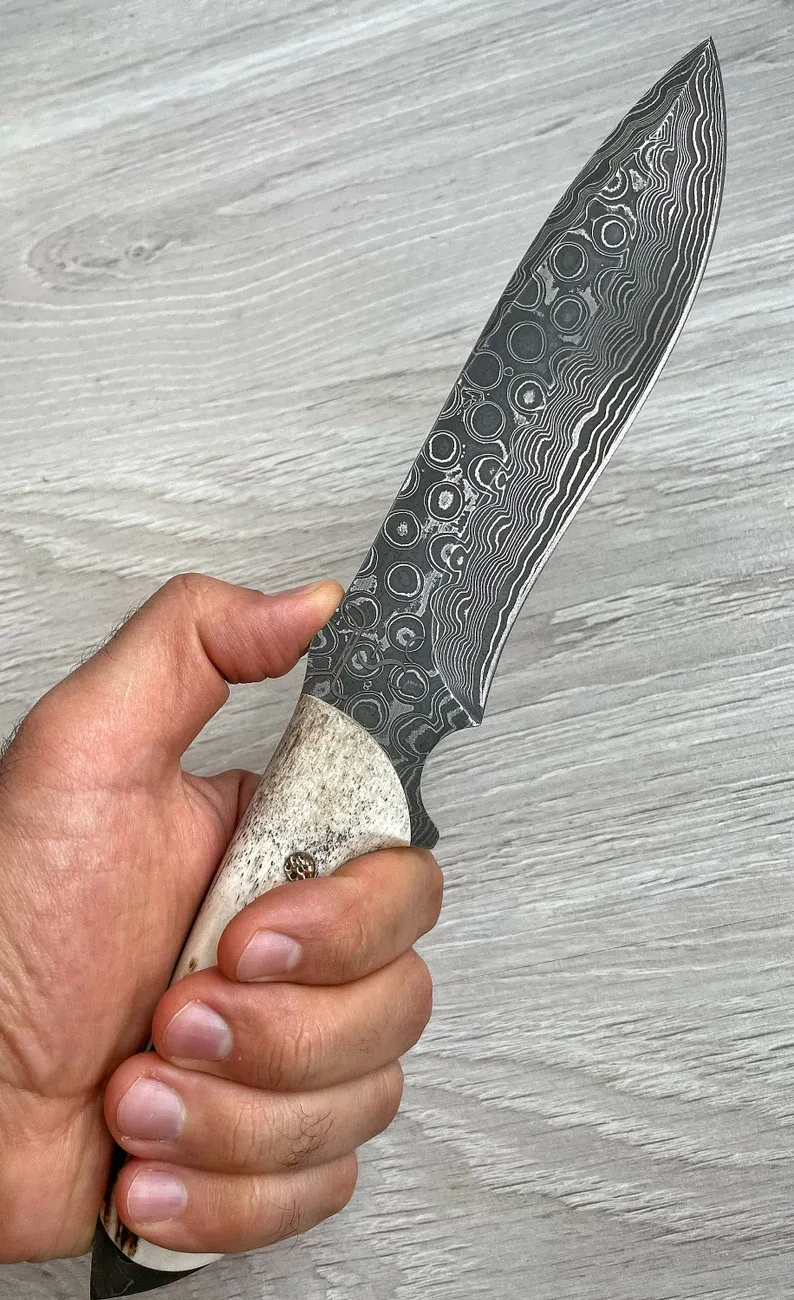Un cuțit Bowie autentic de Damasc, cu m&acirc;ner din corn de cerb și arțar, forjat &icirc;n 150 de straturi, lucrat manual de un fierar.