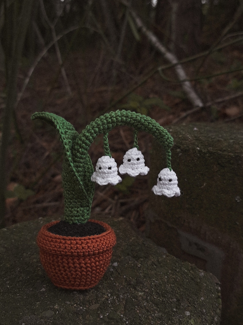 May include: A crocheted lily of the valley plant in a brown pot. The plant has three white flowers with black eyes and green leaves.