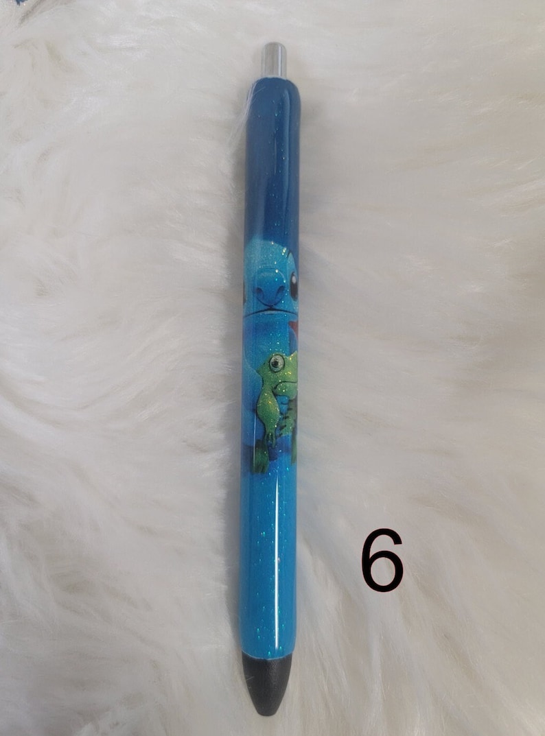 May include: Blue glitter pen with a cartoon image of Stitch and a green alien character. The pen has a black tip and a silver clip. The number 6 is written in black on the white background.
