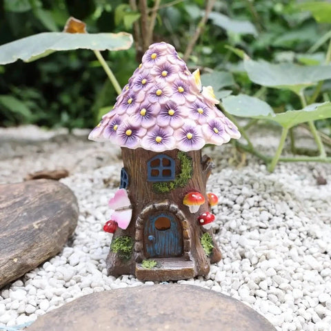A small, decorative fairy house resembling a tree trunk with a purple flower-covered roof sits on white pebbles. It has a blue door and window, and small mushroom and butterfly accents.