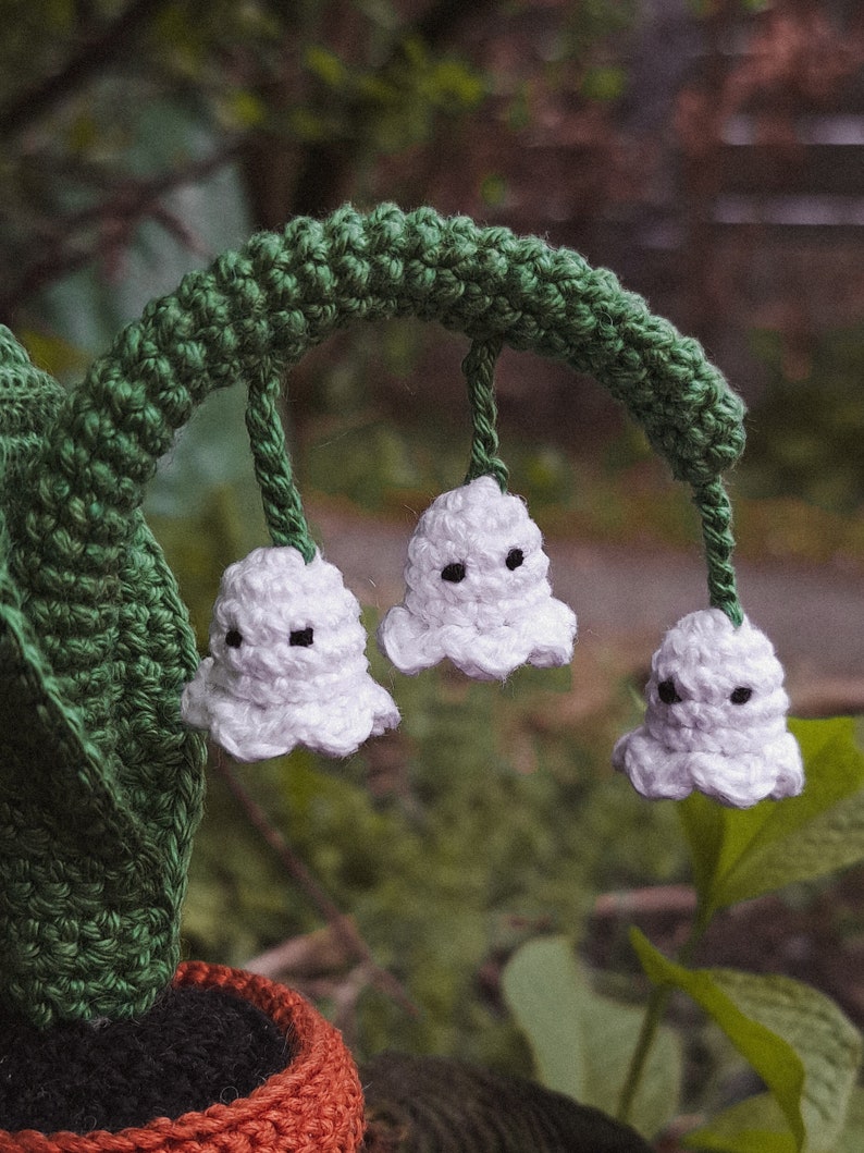 May include: A crocheted lily of the valley plant with three white flowers and green stems. The plant is in a brown and black pot.
