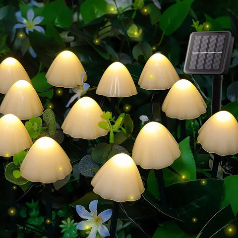 Numerous small, mushroom-shaped solar lights with translucent caps emit a warm white glow among green foliage and small white flowers at night. A small rectangular solar panel on a stake is visible among the lights.