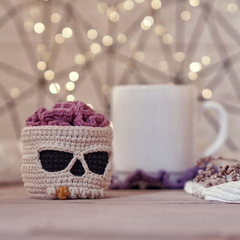 May include: A crocheted skull-shaped basket with a pink brain inside. The basket is beige with black eyes and a beige nose. The basket is sitting on a wooden table in front of a white mug and a blurry background of lights.
