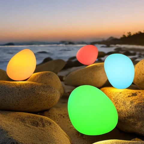 Four glowing, egg-shaped lights of different colors (yellow, red, blue, and green) are scattered across large rocks on a beach at dusk or dawn. The ocean and a colorful sky are visible in the background.