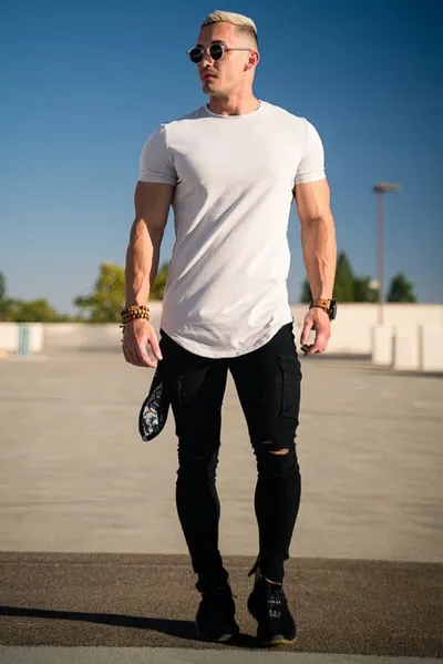 Ein Mann trägt ein weißes T-Shirt, zerrissene schwarze Jeans und eine Sonnenbrille und steht im Freien unter einem klaren blauen Himmel.