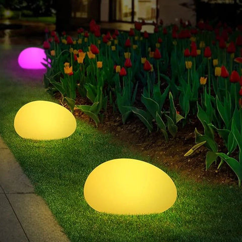 Glowing egg-shaped lights illuminate a garden path at night, with yellow lights on the grass and a purple light among red and yellow flowers. A softly lit building is in the background.