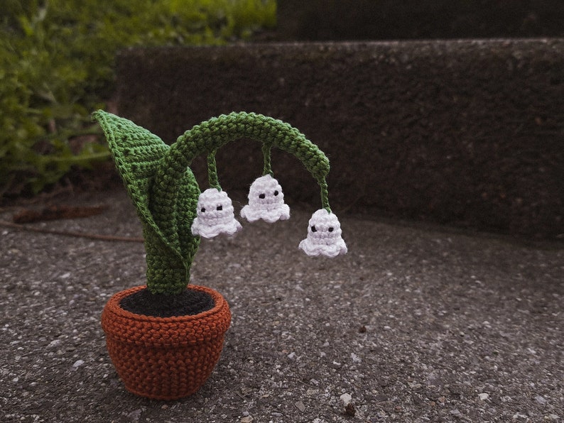 May include: A crocheted plant with green leaves and white flowers shaped like ghosts. The plant is in a brown pot and is sitting on a gray concrete surface.