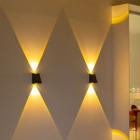 Two small, dark, rectangular lights are mounted on a light-colored wall, each casting a warm white triangular beam both upwards and downwards.