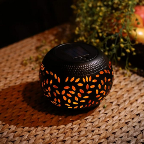 A small, round, black outdoor lantern with decorative oval cutouts emits a warm orange flickering light as it sits on a woven surface. The lantern has a dark, flat top, likely a solar panel. Blurred greenery is visible in the background.