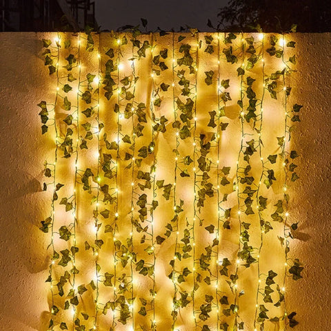 A curtain of warm white string lights intertwined with artificial green ivy leaves hangs against a textured light-colored wall at night. The lights cast a soft glow on the wall behind the foliage.