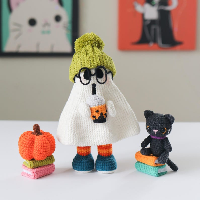 Cute crochet ghost doll with black glasses, green knit beanie, and blue-orange striped boots, sipping a boba tea. Displayed with a stack of colorful crochet books and an orange amigurumi pumpkin. Perfect handmade Halloween or fall decor. This is a PDF digital download crochet pattern to make your own cozy ghost plush and accessories