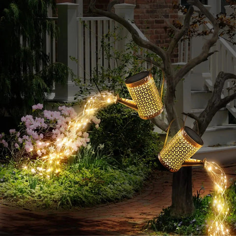 Two decorative, golden watering cans with intricate cutouts are positioned in a garden at dusk or dawn, each illuminated by solar-powered lights within. One watering can is perched on a tree branch, appearing to pour a stream of warm-toned lights onto the greenery below. The other is placed near flowering bushes, with a similar stream of lights extending towards the ground. The illuminated cutouts cast patterns of light, creating a whimsical and enchanting garden scene. A house with white railings is softly lit in the background.