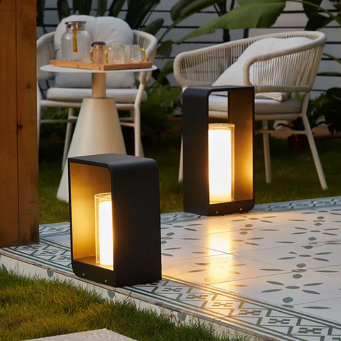 Two modern, dark rectangular outdoor pathway lights with glowing rectangular diffusers, illuminating a tiled patio and grassy area with outdoor furniture.