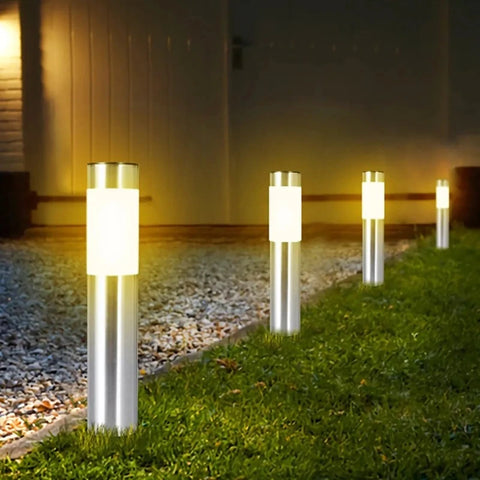 Four cylindrical outdoor pathway lights illuminate a grassy area next to a gravel path at dusk or dawn. The lights have silver metal bases and frosted white diffusers, emitting a warm, upward glow. The lights are spaced out along the edge of the lawn, leading towards a building with a light-colored exterior.