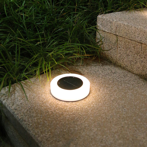 A round, white solar pathway light with a dark center sits on a textured stone surface at night, emitting a bright white upward glow. Green grass grows nearby.