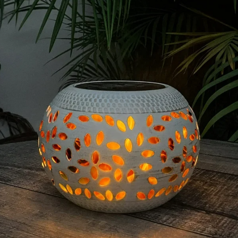 A round, off-white outdoor lantern with decorative oval cutouts emits a warm orange glow as it sits on a dark wooden surface. The lantern has a dark, flat top, likely a solar panel. Palm-like leaves are visible in the blurred background.