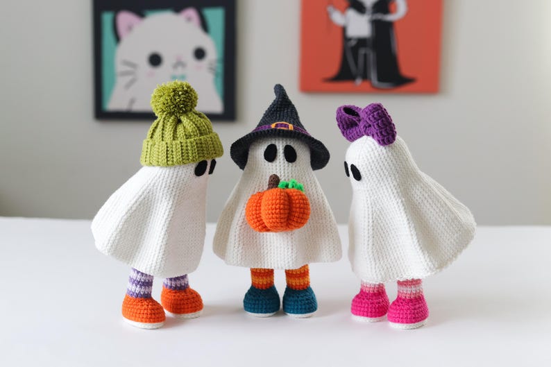 May include: Three crocheted ghost ornaments for Halloween. One ghost wears a witch's hat and holds a small crocheted pumpkin. Another wears a purple crocheted bow, and the third wears a lime green crocheted beanie. All three have white crocheted bodies and colorful crocheted leg warmers and shoes.