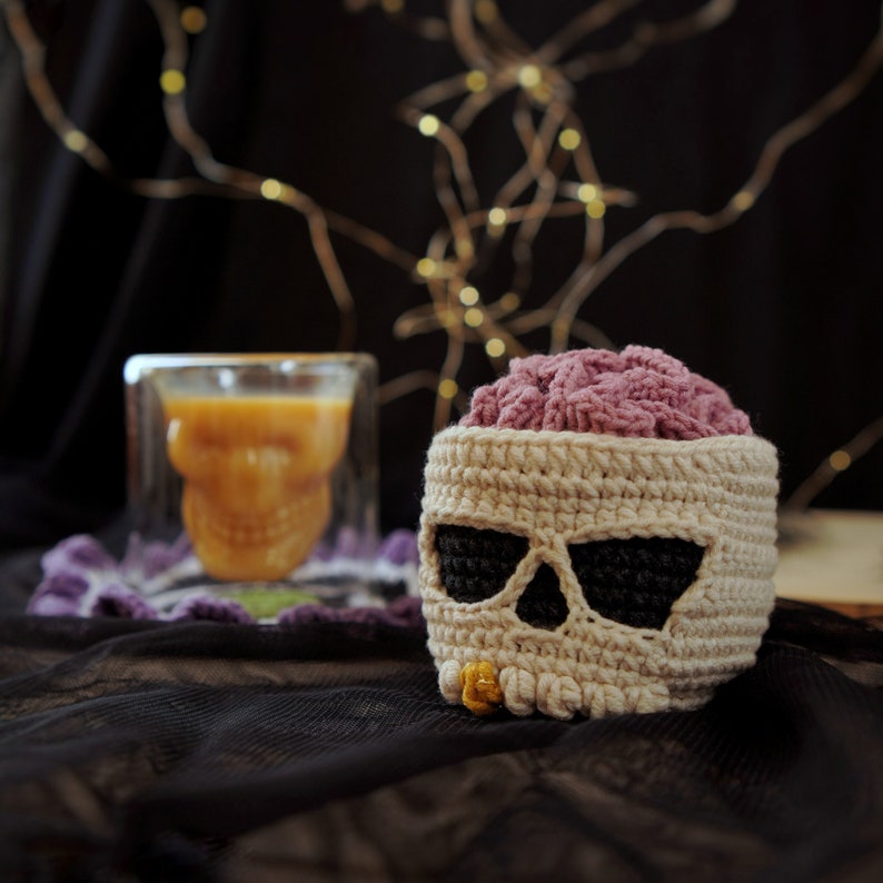 May include: A crocheted skull shaped container with a pink brain inside. The container is made of white yarn with black yarn accents for the eyes and nose. The container is sitting on a black fabric surface.