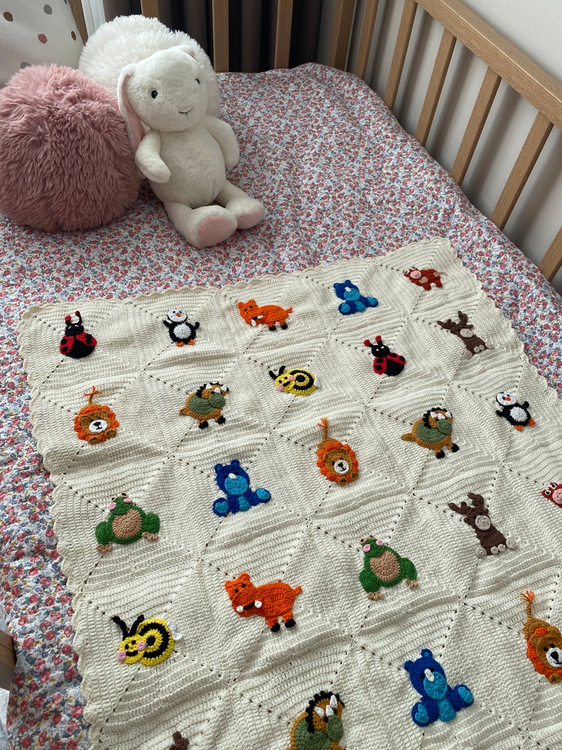 May include: A white crocheted baby blanket with a variety of colorful animal appliques including a lion, a penguin, a ladybug, a bear, a turtle, a frog, a bee, a rhino, and a blue bear. The blanket is laid out on a floral patterned crib sheet.