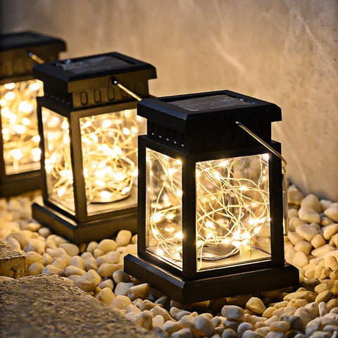 Three solar-powered lanterns with black ABS frames and LED fairy string lights inside, placed on a bed of white pebbles. The lanterns emit a warm, glowing light, creating a cozy and elegant outdoor ambiance. The solar panels on top ensure automatic charging during the day for nighttime illumination.