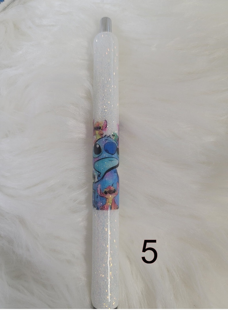 May include: A white glitter pen with a blue and white cartoon character design. The design features a blue character with large eyes and a pink nose. The pen has a silver cap and a black tip. The number 5 is written in black on the white background below the pen.