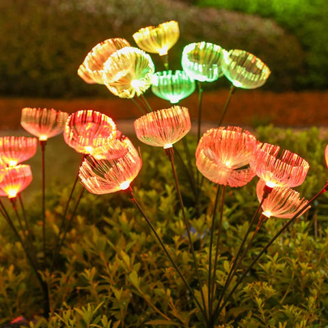 Numerous solar-powered garden lights with a jellyfish-like appearance emit a mix of warm orange, yellow, and green glows in a garden at night, standing above dark green foliage.