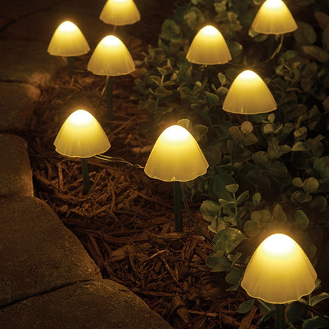 Several small, mushroom-shaped solar lights with translucent caps emit a warm yellow glow in a garden at night. The lights are on thin stakes and are scattered among rocks, mulch, and small plants.