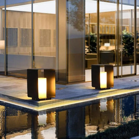 Two modern, dark square outdoor pathway lights with glowing cylindrical diffusers, illuminating a walkway alongside a reflecting pool near a building with large glass windows.