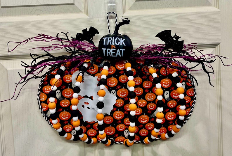 可能包含:一个带有黑色和橙色南瓜图案的万圣节花环。花环上装饰着黑白珠子、橙白珠子和黑色蝙蝠。花环中央有一个黑色南瓜,上面写着“不给糖就捣蛋”的字样。